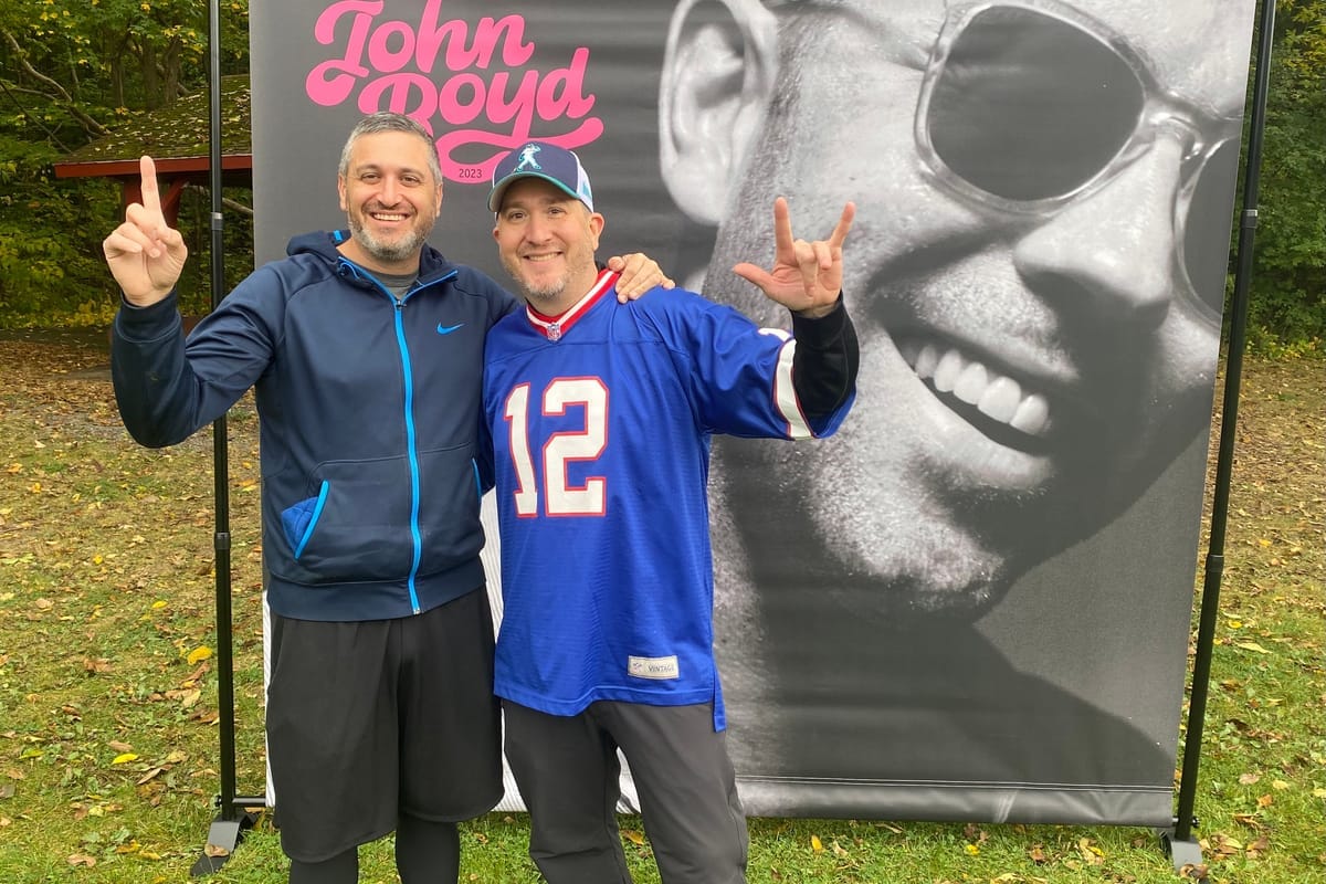 Two friends in Bills gear posing in front of the banner of John laughing