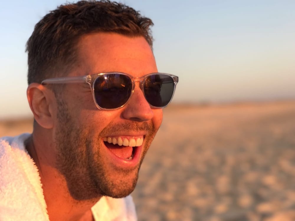 John Boyd laughing in golden evening light on a beach, wearing sunglasses