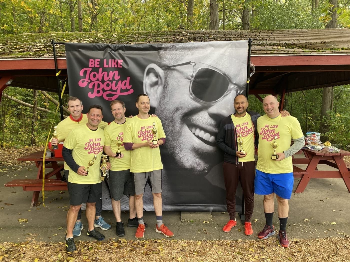 Six players in yellow Be Like John Boyd shirts holding trophies in front of a banner of John laughing